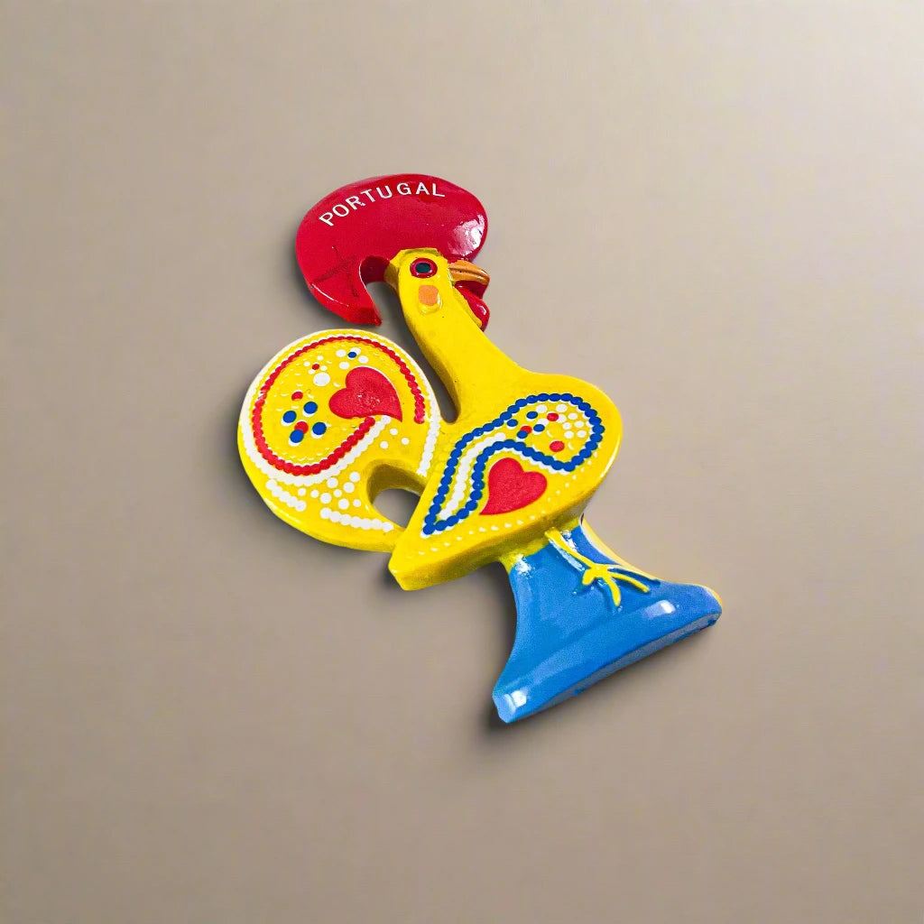 A vibrant ceramic rooster figurine adorned with detailed red, blue, and yellow patterns, mounted on a blue base. The rooster features a red comb and wattle, with "Portugal" inscribed on its comb, symbolizing the Galo de Barcelos. This delightful piece also serves as a magnet souvenir.