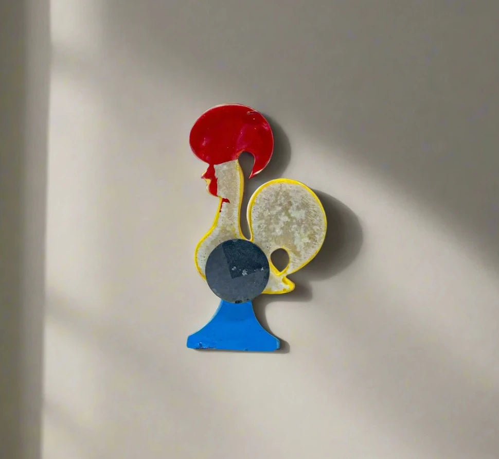 A vibrant ceramic rooster figurine adorned with detailed red, blue, and yellow patterns, mounted on a blue base. The rooster features a red comb and wattle, with "Portugal" inscribed on its comb, symbolizing the Galo de Barcelos. This delightful piece also serves as a magnet souvenir.