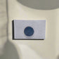 A small white rectangular card, perhaps a unique souvenir, is centered against a neutral background. The card features a textured surface with a dark blue circle in its middle. Shadows from an unseen source are cast around the card, adding depth and contrast to the composition.