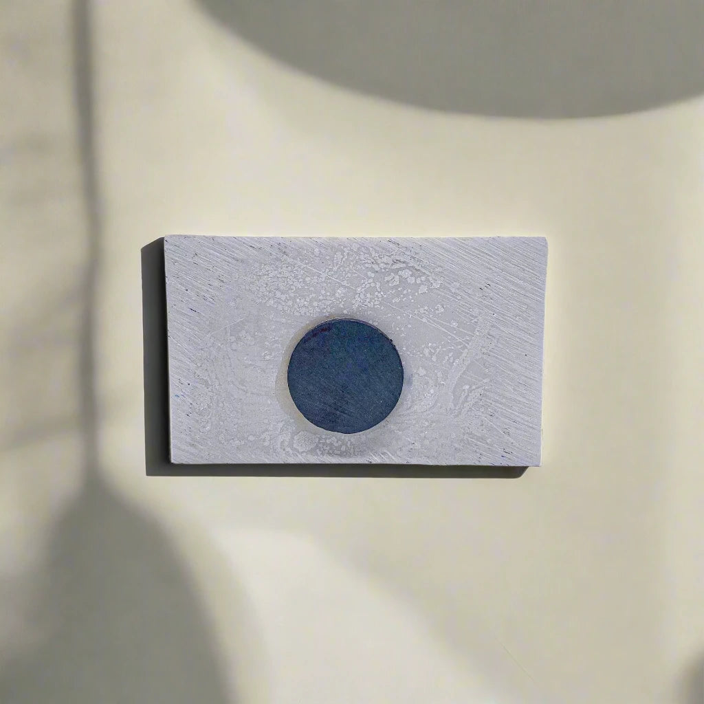 A small white rectangular card, perhaps a unique souvenir, is centered against a neutral background. The card features a textured surface with a dark blue circle in its middle. Shadows from an unseen source are cast around the card, adding depth and contrast to the composition.