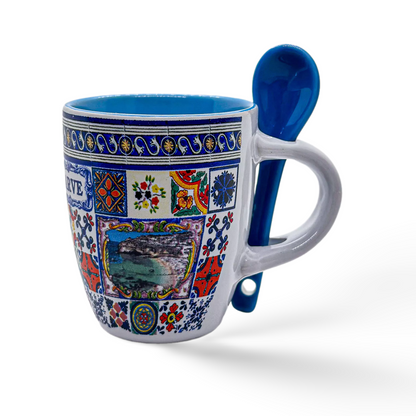 A decorative ceramic coffee cup with colorful, intricate patterns and a small scenic picture at the center. The mug has a built-in slot on the handle holding a matching blue ceramic spoon. The inside of the mug is also blue. This beautiful souvenir from Algarve stands out against its white background.