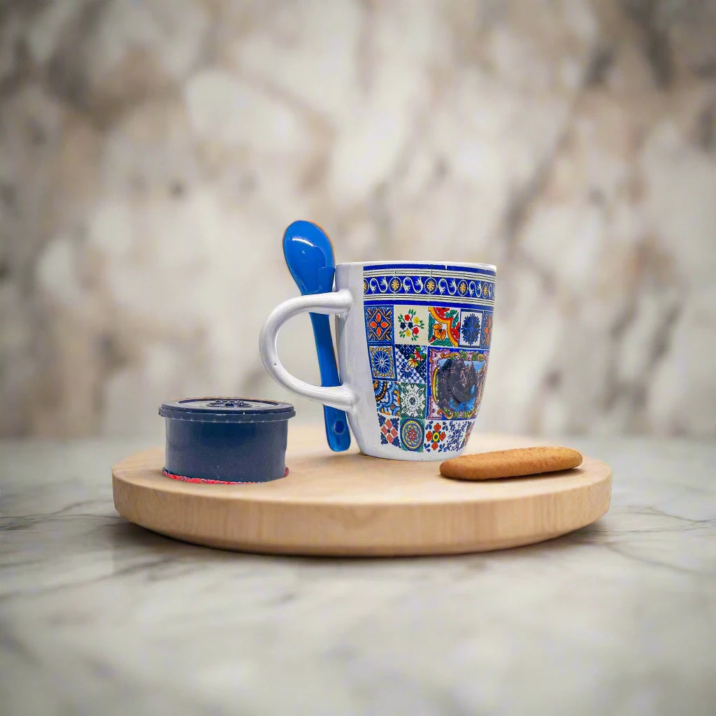 A ceramic coffee cup adorned with vibrant, intricate patterns and a small scenic image in the center. The handle includes a built-in slot holding a matching blue ceramic spoon, and the inside of the cup is also blue. This stunning Algarve souvenir stands out against its white backdrop.