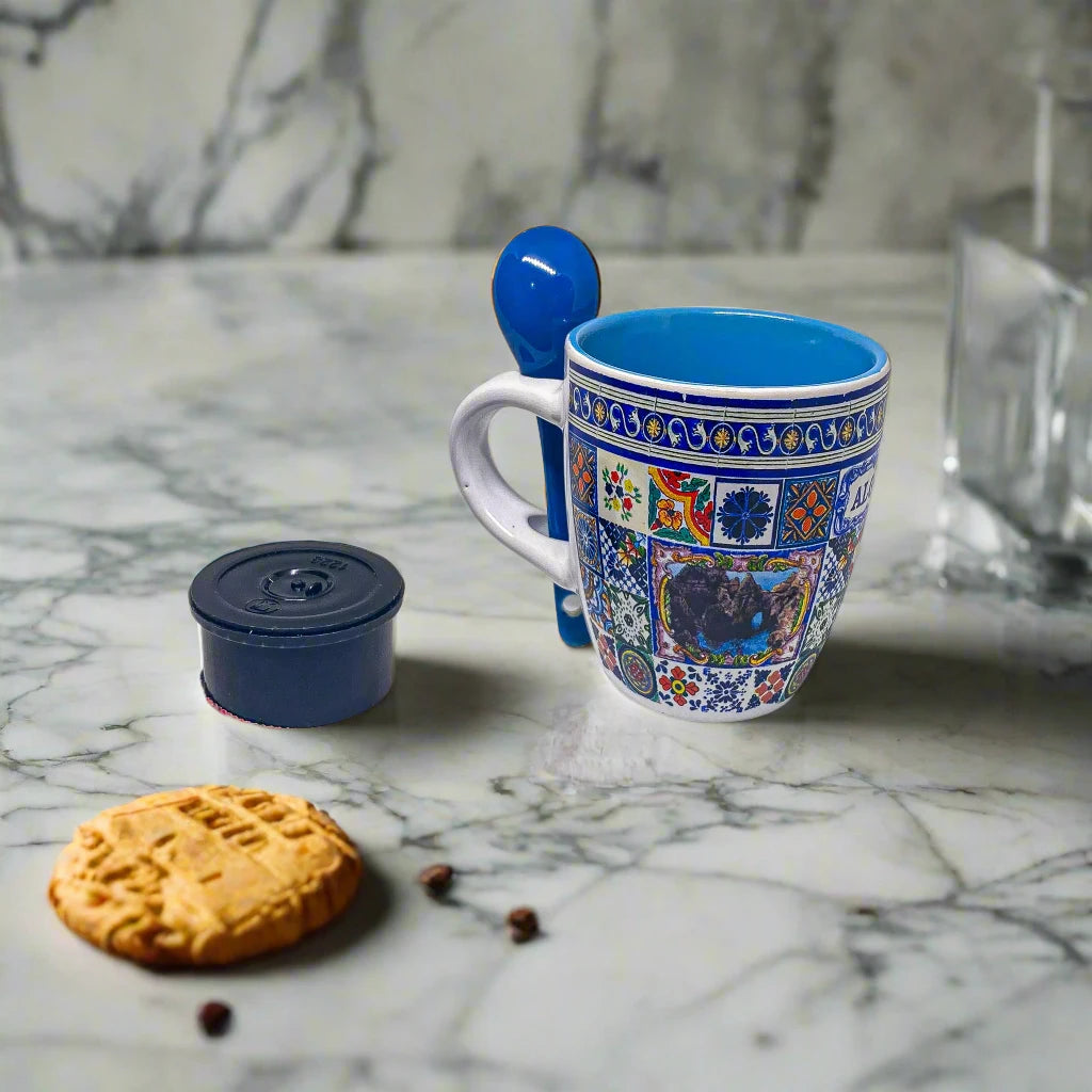 A beautifully designed ceramic coffee cup with intricate, colorful patterns and a small scenic image in the middle. The handle has a slot for a matching blue ceramic spoon, and the mug's interior is also blue. This eye-catching Algarve keepsake stands out against its white surface.