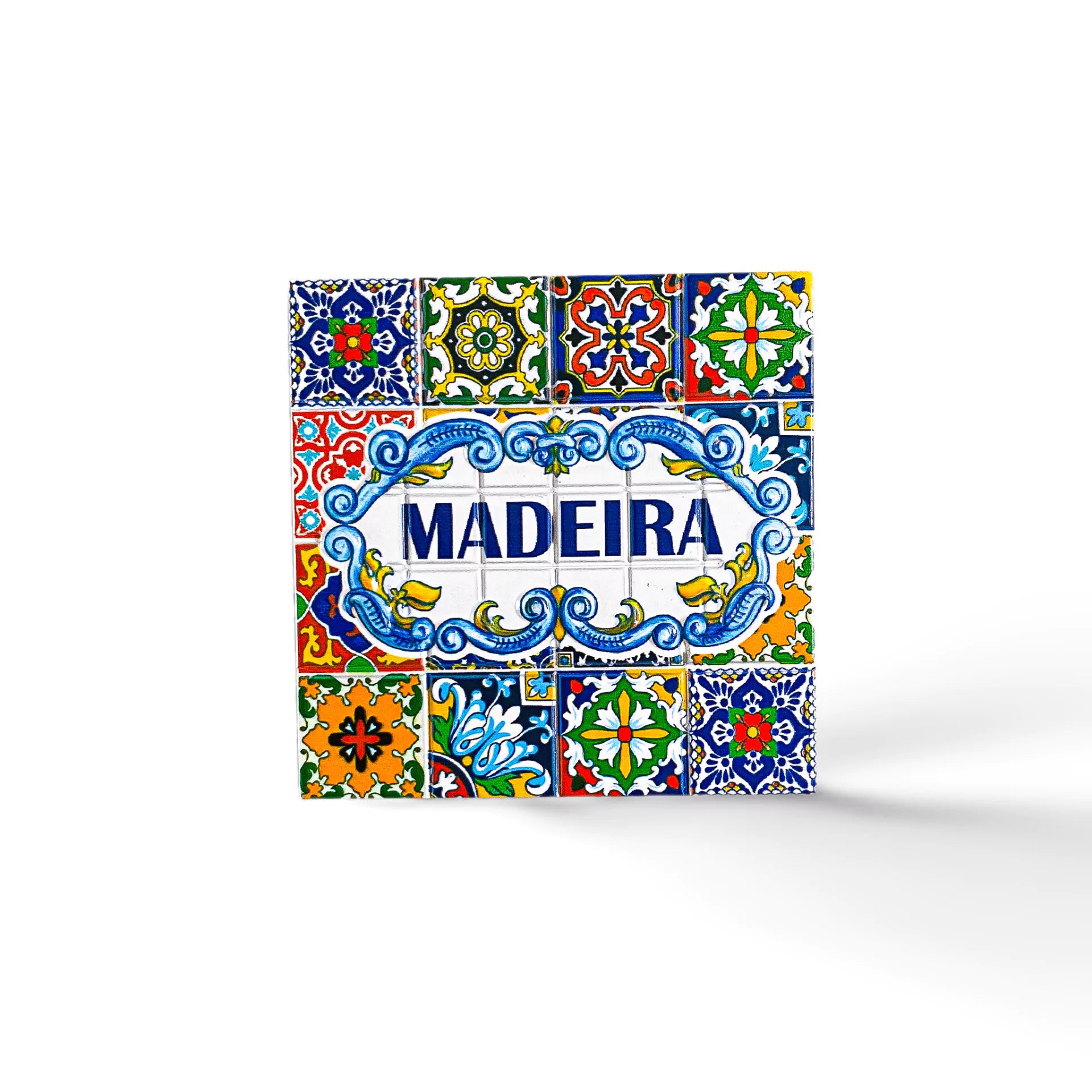 A decorative square tile, perfect as a souvenir from Portugal, showcases the word "Madeira" in blue text at its center, surrounded by intricate, colorful patterns and floral designs in shades of yellow, green, red, and blue. The white background highlights the vibrant details of this charming magnet.