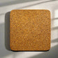 A square corkboard with rounded edges is mounted on a light-colored wall. Shadows from slatted blinds fall across the surface, creating parallel lines of shade. The corkboard, sourced from Portugal, has a textured surface with varying tones of brown.