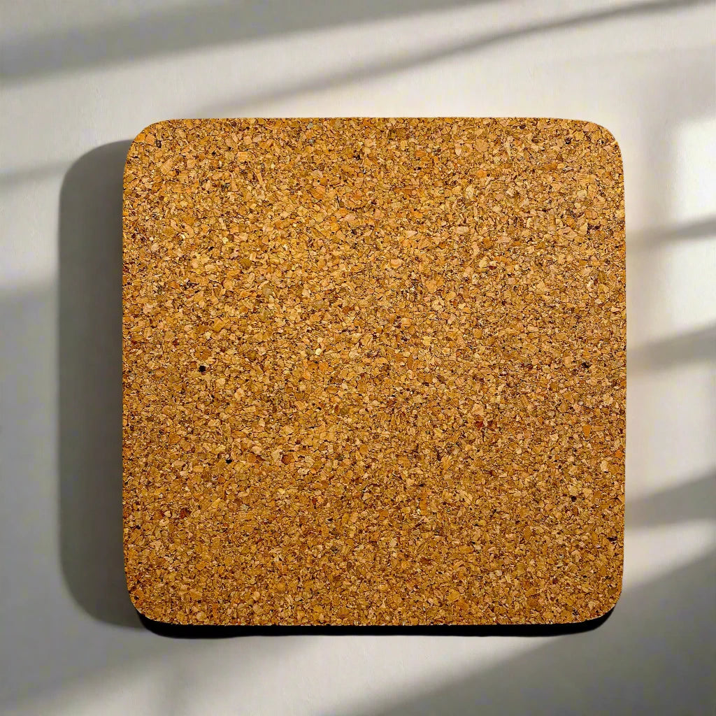 A square corkboard with rounded edges is mounted on a light-colored wall. Shadows from slatted blinds fall across the surface, creating parallel lines of shade. The corkboard, sourced from Portugal, has a textured surface with varying tones of brown.
