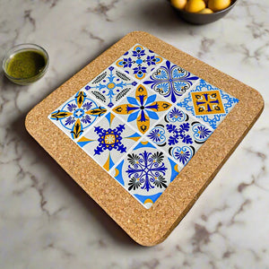 A square tile coaster featuring intricate blue, yellow, and white patterns, including floral and geometric designs arranged in a 3x3 grid. This delightful Portuguese souvenir has a sturdy cork base and is set on a white surface.