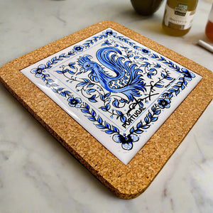 A square cork coaster with a blue and white ceramic tile at the center, showcasing a stylized rooster surrounded by intricate floral patterns. The word "Portugal" is displayed at the bottom, making it a charming souvenir from Portugal.