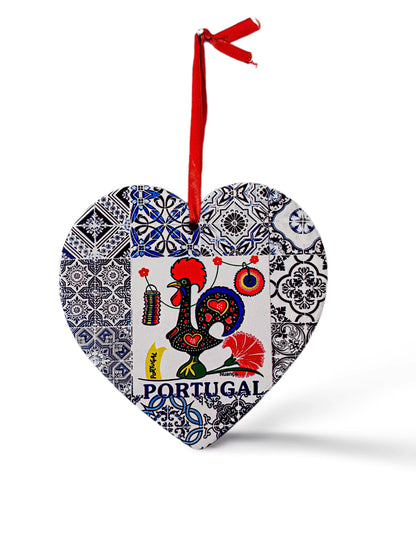 A heart-shaped ornament features a colorful rooster design with the word "Portugal" written below it. The rooster is surrounded by intricate blue and white tile patterns. A red ribbon is attached to the top for hanging, and a cork base adds a charming touch to this delightful souvenir.