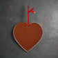 A heart-shaped cork board, sourced as a cherished souvenir from Portugal, hangs against a dark, textured wall. Suspended by a red ribbon tied through a hole at the top of the heart, it features a small jingle bell attached to the ribbon, adding a decorative touch.