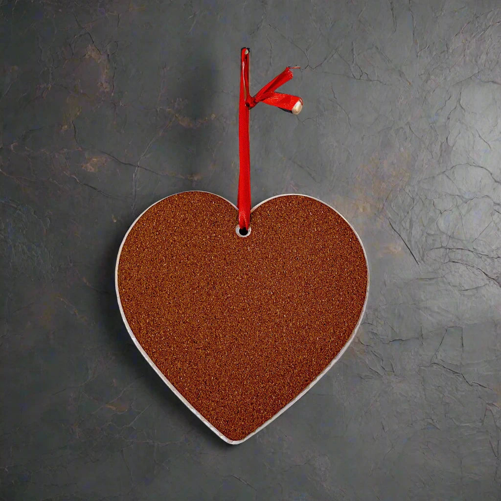A heart-shaped cork board, sourced as a cherished souvenir from Portugal, hangs against a dark, textured wall. Suspended by a red ribbon tied through a hole at the top of the heart, it features a small jingle bell attached to the ribbon, adding a decorative touch.
