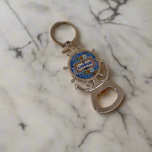 A silver keychain bottle opener, designed in the shape of a ship's wheel and anchor, is adorned with a blue and yellow mosaic pattern and the word "Lisboa" at the center. This delightful keepsake is ideal for any fan of Lisbon.