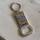 A metal keychain with a bottle opener at the base, topped by a rectangular enamel tile showcasing a depiction of a Lisbon tram. The word "Lisboa" is displayed above the tram, framed by a decorative blue and yellow border, encapsulating the charm of Portugal.