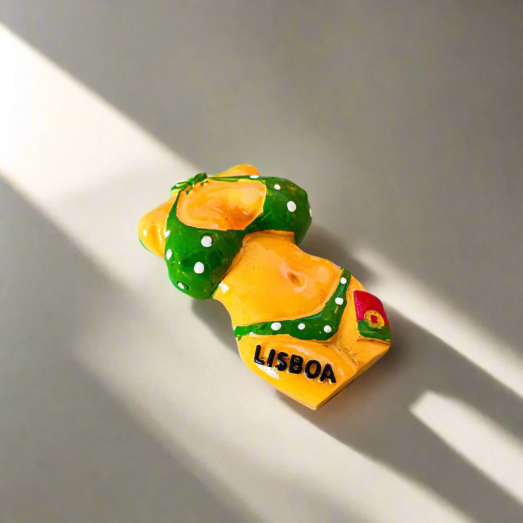 A small magnet showcasing a figurine of a bikini-clad girl in a green bikini with white polka dots. The word "LISBOA" is printed on the left side of the bottoms, with a tiny Portuguese flag on the right. This cute souvenir contrasts nicely with the white background.