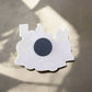 A decorative white train-shaped souvenir magnet from Portugal is adhered to a metallic surface, partially illuminated by soft, diffused sunlight. The magnet has rounded and angular edges, representing a classic steam locomotive silhouette with a prominent black circle in the center.