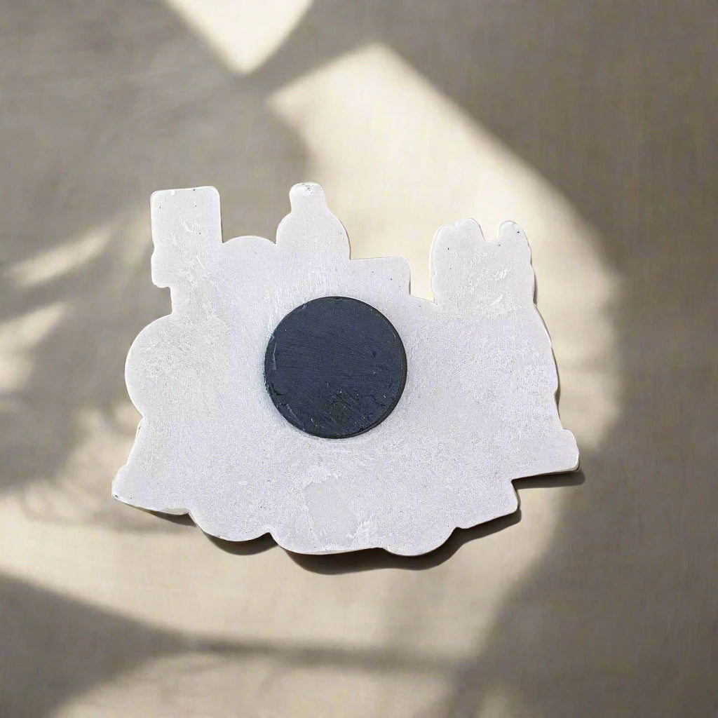 A decorative white train-shaped souvenir magnet from Portugal is adhered to a metallic surface, partially illuminated by soft, diffused sunlight. The magnet has rounded and angular edges, representing a classic steam locomotive silhouette with a prominent black circle in the center.
