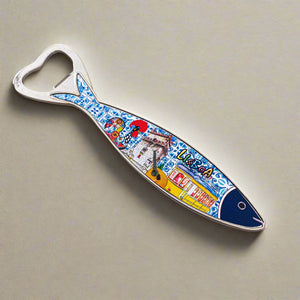 A bottle opener shaped like a sardine, featuring a vibrant illustration of Lisbon, Portugal on the handle. The artwork showcases iconic city landmarks, traditional Portuguese tiles, and a tram. The elongated handle leads to the opener at the fish's mouth, and it also serves as a magnet for your fridge.