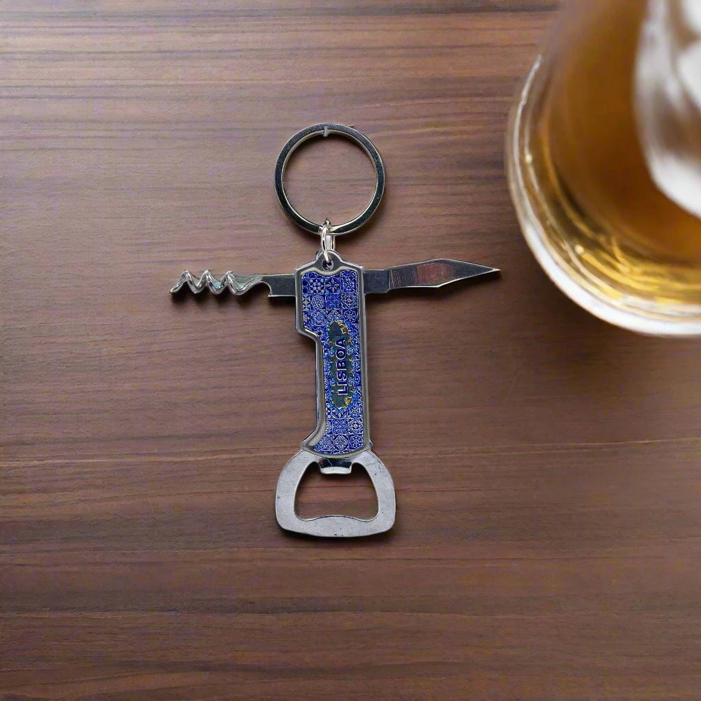 A multipurpose metal keychain shaped like the number 1 rests on a wooden surface. It incorporates a corkscrew, a small knife, and a bottle opener. A glass of beer is partially visible on the right edge of the image, making it the perfect souvenir from Portugal.