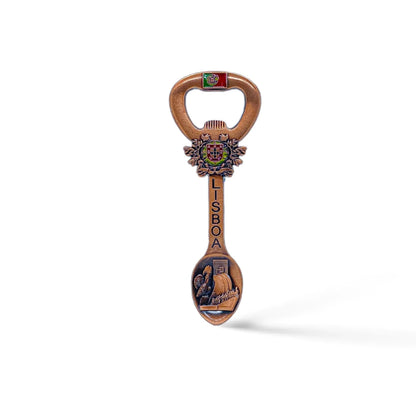 A decorative bottle opener shaped like a spoon, featuring the word "LISBOA" and ornate designs, including a crested emblem and a detailed scene on the spoon part. The handle displays the flag of Portugal. It also has a magnet for easy display on your fridge. The background is white.