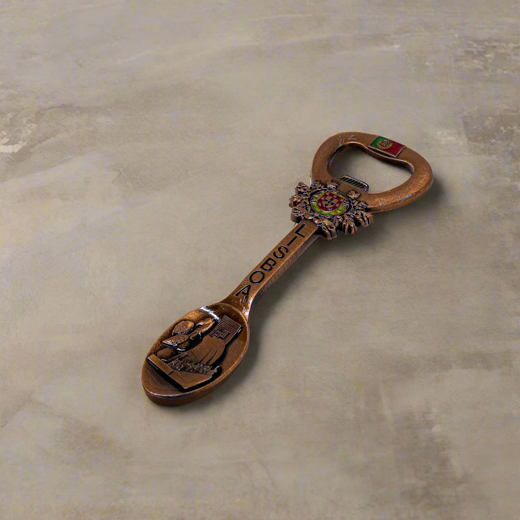 A decorative bottle opener shaped like a spoon, adorned with the word "LISBOA" and intricate designs, including a crested emblem and a detailed scene on the spoon bowl. The handle showcases the Portuguese flag, and a magnet on the back allows for easy fridge display. 