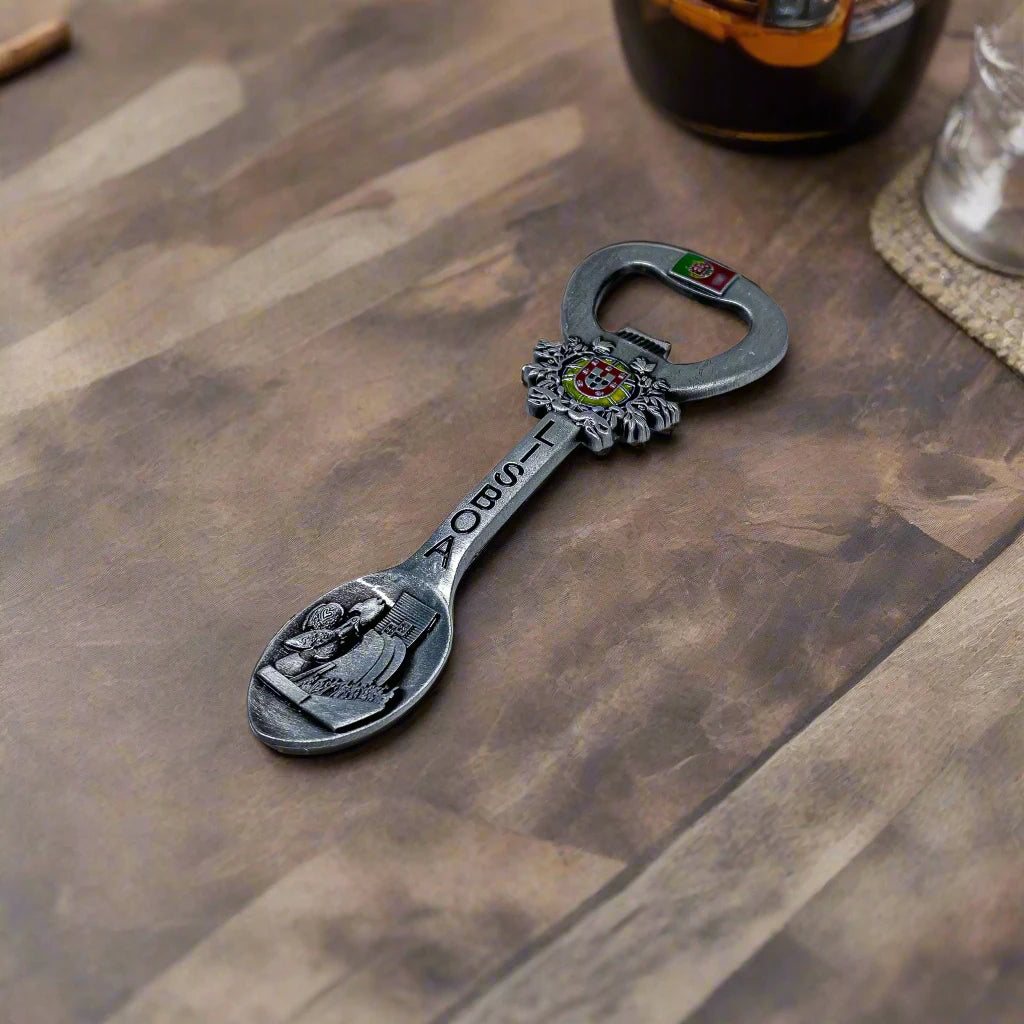 A versatile souvenir from Lisbon, Portugal, combining a bottle opener, magnet, and spoon. The piece is adorned with the Portuguese flag, the Lisbon crest, the word "Lisboa," and an engraved depiction of a famous Lisbon landmark on the spoon end.