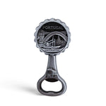 A silver bottle opener souvenir featuring an engraved image of a bridge and cityscape labeled "PORTUGAL" with "PORTO-PORTUGAL" written underneath. The handle also has an engraving of a tram. The background is a plain white.