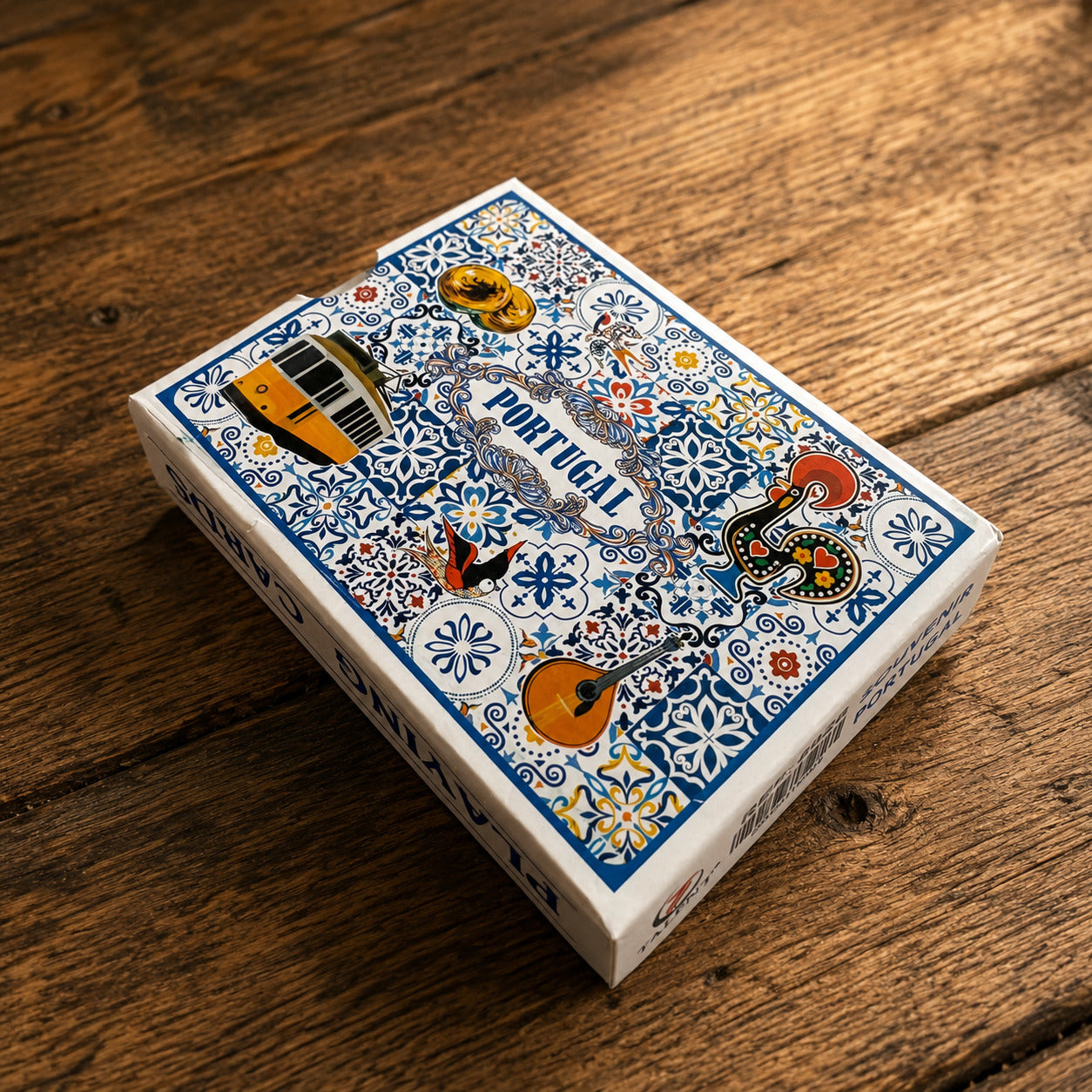 Portugal souvenir playing cards box displayed on a rustic wooden surface, decorated with traditional azulejo motifs, yellow tram, Barcelos rooster, Portuguese guitar, and cultural illustrations.