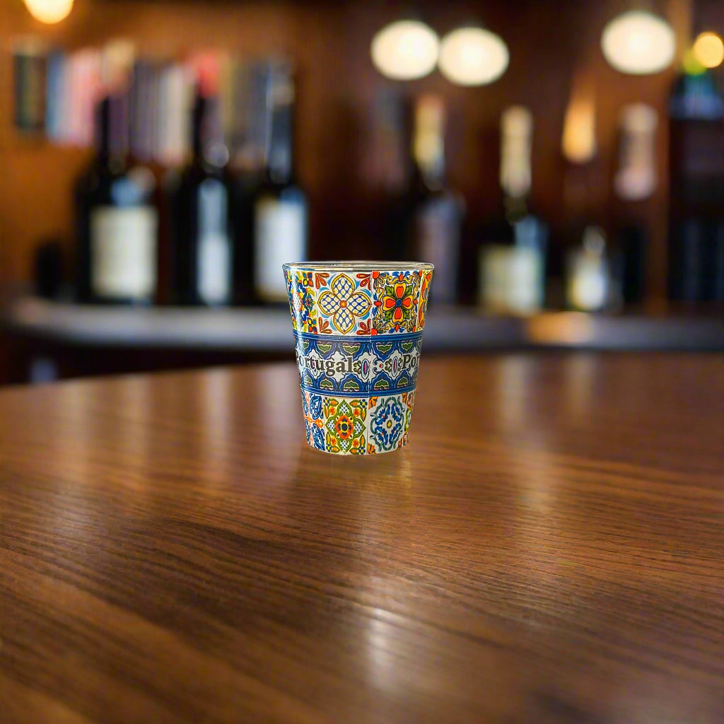 Portugal shot glass featuring vibrant Portuguese tile artwork displayed on a wooden bar counter with blurred bottles in the background.