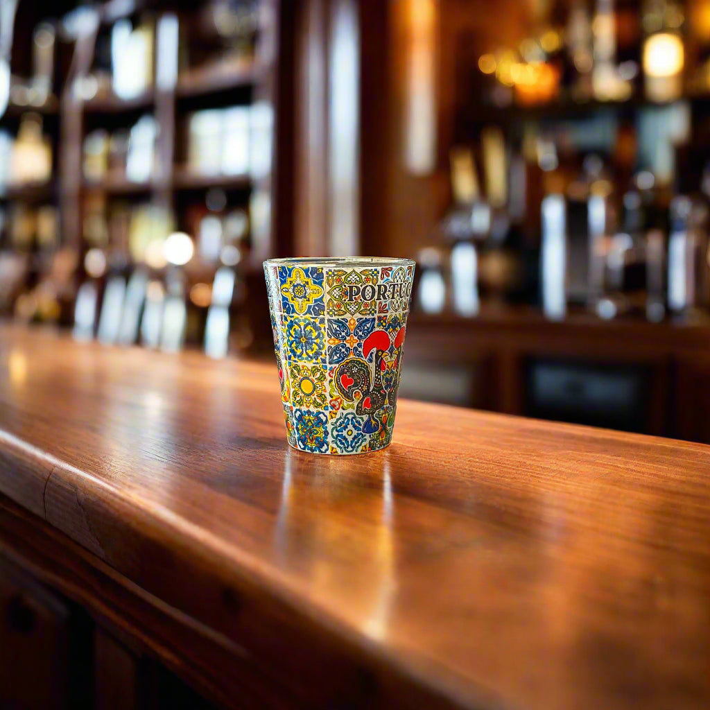 Colorful Portugal souvenir shot glass featuring Barcelos rooster illustrations and classic Portuguese tile patterns on a rustic wooden bar.