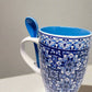 This ceramic coffee mug, adorned with blue and white traditional Portuguese tile motifs, features the word "PORTUGAL" around its base. It includes a coordinating blue spoon that fits neatly into a slot on the handle, combining elegance with practicality.
