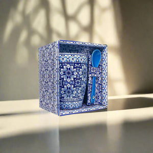 
A beautifully detailed ceramic coffee mug with intricate blue and white patterns, reminiscent of traditional Portuguese tile art, is packaged in a matching decorative box. This charming souvenir includes a blue ceramic spoon resting on the side of the mug, secured within the box. Shadows from a window's pattern fall across the background.