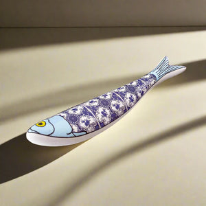 A ceramic sardine figurine adorned with a blue and white intricate pattern, inspired by traditional Portuguese tiles. The fish features a blue head with a yellow eye, and its body is segmented into four sections with the detailed pattern. The word "Portugal" is elegantly placed near the tail, making it an ideal souvenir.