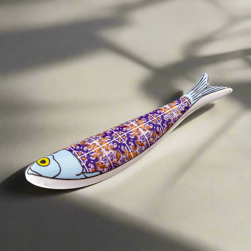 A tiny ceramic fish shaped like a sardine, decorated with detailed purple and orange tile patterns, featuring a light blue head and a yellow eye. 