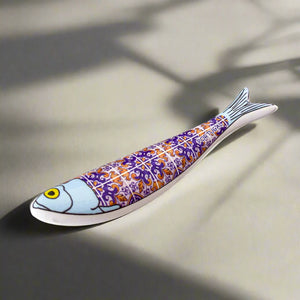 A tiny ceramic fish shaped like a sardine, decorated with detailed purple and orange tile patterns, featuring a light blue head and a yellow eye. 