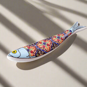 A decorative ceramic fish magnet with a blue head, yellow eye, and a detailed, colorful body pattern that resembles traditional Portuguese tiles. The body is adorned with vibrant reds, blues, and yellows, with the word "Portugal" displayed near the tail. 
