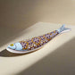 A stylized fish sculpture, inspired by the iconic Portuguese sardine, showcases a blue head and a body adorned with intricate patterns in yellow, white, and purple. The fish has a prominent yellow eye and a light blue tail that subtly blends into the background, making this vibrant piece a striking magnet as well as a beautiful decorative item.