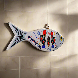 A fish-shaped souvenir wall decoration with "Portugal" inscribed on its side, showcasing three stylized roosters decorated in vibrant red, yellow, and blue details. The fish features a bright yellow eye and has a triangular hanger at the top, making it a striking and colorful addition to any space.