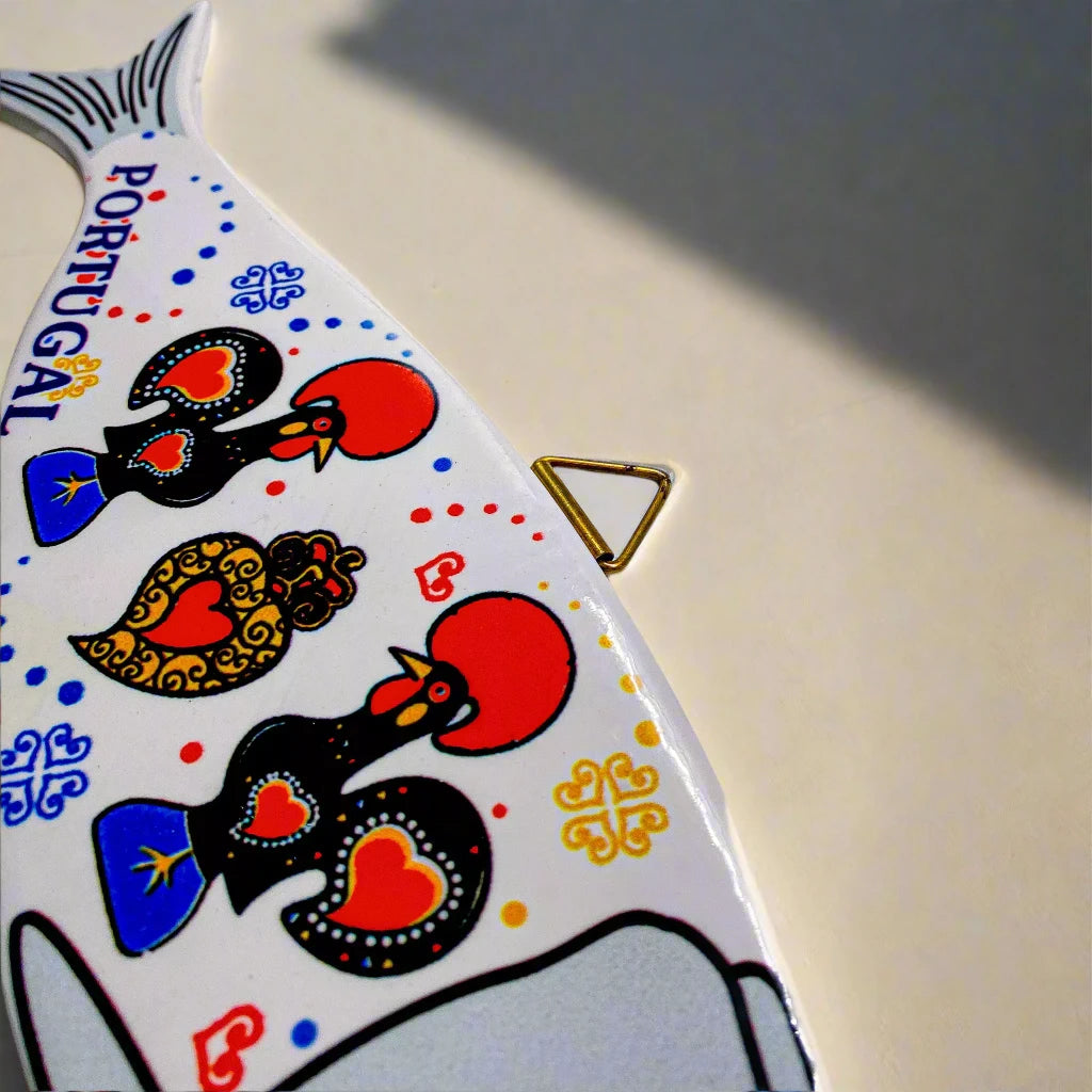 A fish-shaped wall decoration, with "Portugal" prominently displayed along its side, features three intricately designed roosters in bold red, yellow, and blue accents. The fish has a yellow eye and a triangular hanger at the top, making it a vibrant and eye-catching piece perfect for adding a touch of Portuguese charm to any room.