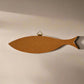 A corkboard shaped like a sardine is mounted on a plain wall. The sardine corkboard has a small brass hanger attached at the top for easy wall mounting. The overall look is minimalist with a focus on the unique fish shape and its cork base.