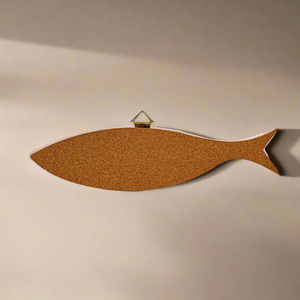 A corkboard shaped like a sardine is mounted on a plain wall. The sardine corkboard has a small brass hanger attached at the top for easy wall mounting. The overall look is minimalist with a focus on the unique fish shape and its cork base.