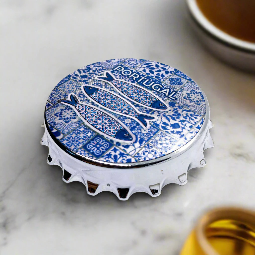 A bottle cap design featuring three fish with intricate patterns set against a blue and white tiled background, reminiscent of traditional Portuguese tiles. The word "Portugal" is prominently placed above the fish, while the cap's classic crimped edges add a touch of authenticity. It also doubles as a magnet, blending functionality with style.