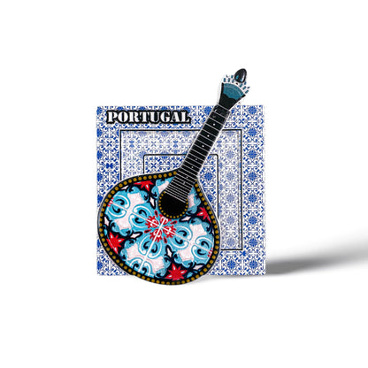A magnet featuring the word "Portugal" at the top, with a traditional Portuguese "guitarra classica" at its center. The guitar showcases an ornate, colorful design with blue, red, and black patterns. The backdrop consists of a white square adorned with intricate blue tile patterns.