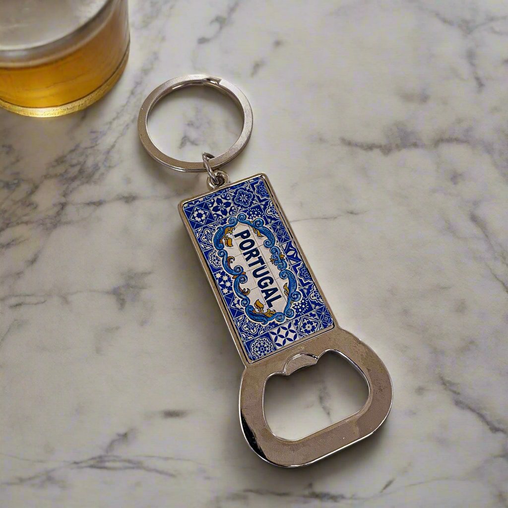 A rectangular keychain with a built-in bottle opener, featuring detailed blue patterns and "Portugal" displayed prominently in the center. The metallic ring complements the blue and silver-toned opener, creating a striking contrast. This elegant keepsake also doubles as a magnet, making it an ideal souvenir for any travel enthusiast.