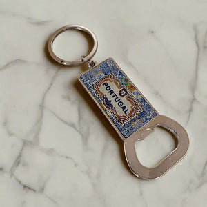 A souvenir keychain featuring a vibrant blue and multicolored enamel rectangular pendant with intricate designs and the word "PORTUGAL" in bold lettering. A convenient bottle opener is attached at the bottom, while the keychain ring is sleek and black, adding a stylish touch to this functional keepsake.