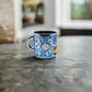 A small metal shot glass, resembling a shot glass, with a blue and white intricate pattern sits on a smooth, stone-like surface. The cup features abstract designs, including a circle, and is slightly reflective. This charming piece makes for a perfect souvenir from Portugal. The background is blurred, highlighting the cup as the focal point.