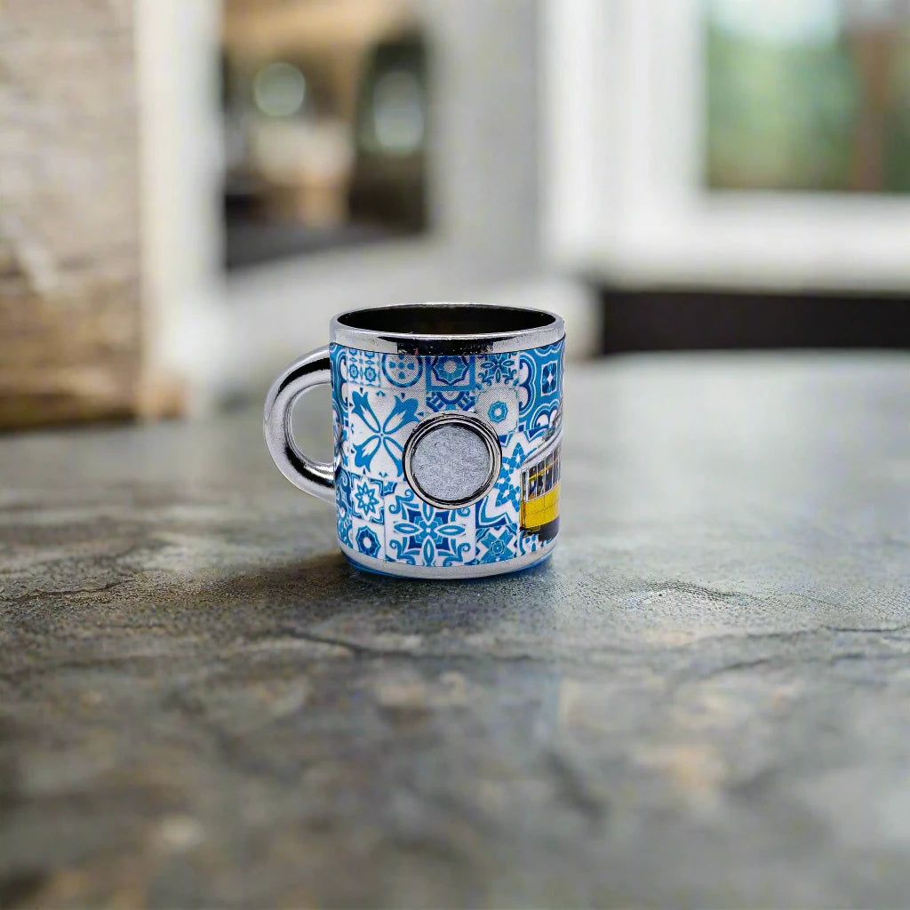 A small metal shot glass, resembling a shot glass, with a blue and white intricate pattern sits on a smooth, stone-like surface. The cup features abstract designs, including a circle, and is slightly reflective. This charming piece makes for a perfect souvenir from Portugal. The background is blurred, highlighting the cup as the focal point.