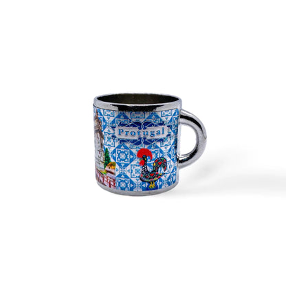 A small metal mug features the word "Portugal" on a blue and white patterned background reminiscent of traditional Portuguese tiles. The design includes colorful illustrations, such as the iconic Portuguese rooster, making it a delightful addition next to your favorite Portugal-themed magnets.