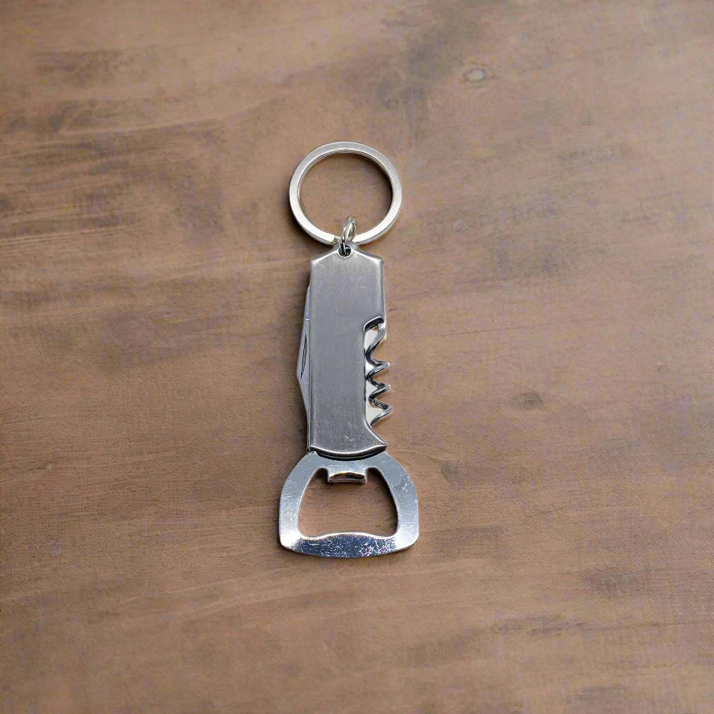 A compact, multi-functional keychain rests against a wooden backdrop. This metal tool from Portugal features a knife blade, corkscrew, and bottle opener, with a keyring attached at the top for easy carrying.