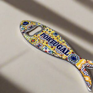 A fish-shaped metal bottle opener adorned with intricate and colorful patterns stands out against a plain background. The word "PORTUGAL" is prominently displayed in blue on the sardine's body. This delightful souvenir doubles as a magnet, making it an ideal keepsake for decorating your fridge.