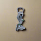 A metallic bottle opener shaped like a Portuguese girl kneeling with her hands behind her head. She is wearing a bikini decorated with the colors and emblem of Portugal, with "Portugal" written at the top. The opener also includes a magnet on the back, allowing for convenient storage on any metal surface.
